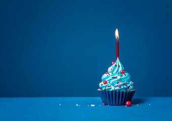 Blue cupcake with red sprinkles and lit candle on a blue background.