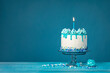 White birthday drip cake with teal ganache and lit candle over blue background