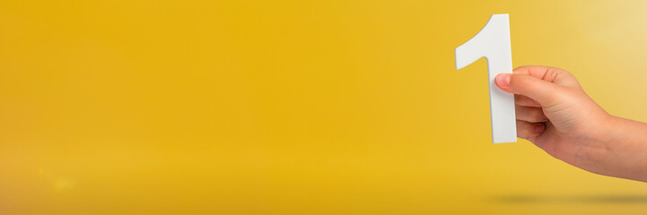 Fototapete - Number one in hand. A hand holds a white number one on a yellow background with copy space. Concept with number one. 1 percent rate, birthday, first or winner.
