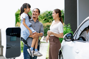 Progressive young parent teach daughter how to recharge or refuel EV car at home charging station. Green and clean energy from electric vehicle for healthy environment. Eco power from renewable source