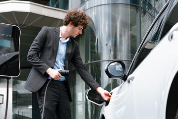 Below view closeup image of progressive black suit businessman recharge battery of his electric vehicle from public charging station. Renewable and alternative energy powered car concept.