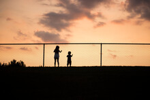 Rear View Of Siblings Standing By Chainlink Fence Against Cloudy Sky During Sunset