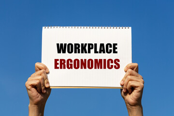 workplace ergonomics text on notebook paper held by 2 hands with isolated blue sky background. this 