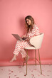 beautiful woman in heart print pajamas on a pink background is working on a laptop. Pink pajamas with red hearts. clothes for sleep and home. Valentine's Day. work at the computer in pajamas at home