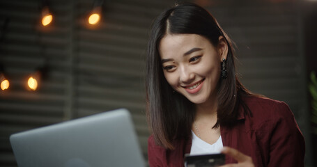 Young asian girl sitting on her bed with laptop, having online shopping, keying in credit card. Asian student making purchase via internet - closeup 