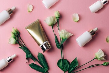 Wall Mural - White cosmetic serum bottles, golden cream tube and eustoma flowers on pink background. Top view, flat lay