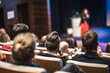 © kasto - Business and entrepreneurship symposium. Female speaker giving a talk at business meeting. Audience in conference hall. Rear view of unrecognized participant in audience.