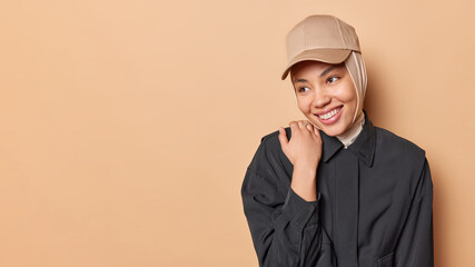 Wall Mural - Happy shy woman smiles gently keeps hand on shoulder looks somewhere aside wears hijab black jacket and cap isolated over brown background copy space for your advertising content. Stylish Muslim girl