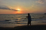 Fototapeta  - An amateur old fisherman on the beach at a scarlet sunset throws his fishing rod into the sea