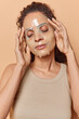 © WHstudio Leushin N - Calm young woman applies collagen face mask for cleansing pores of skin keeps eyes closed dressed casually undergoes daily beauty treatments isolated over brown background. Skin care concept