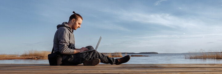 Young male working online on a laptop while sitting on a wooden pier. Freelancer using laptop for internet work outside. The concept of remote work. Banner for website header design with copy space.