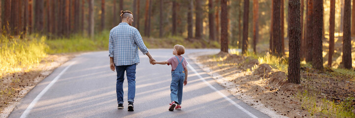 Dad and his little daughter are walking along a forest road among tall pines. Family walk in the forest at sunset, man and little girl. Banner for website header design with copy space.