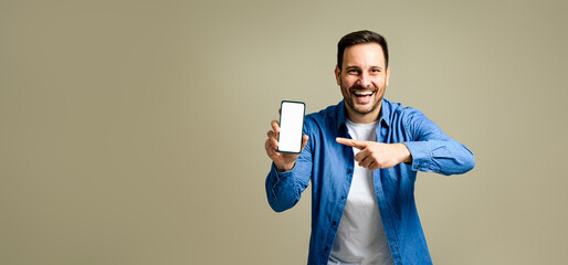 Handsome mid adult man dressed in denim jacket laughing and pointing at smart phone's blank screen isolated against gray background