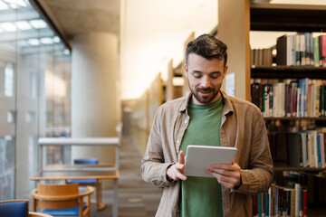 Bearded middle aged man is using digital tablet, looking for e book. Attractive latin student in library, concept of online education, studying at university, college 