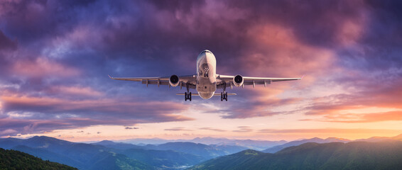 Plane is flying in colorful sky at sunset. Landscape with passenger airplane, mountains, purple sky with orange and pink clouds. Aircraft is landing. Business travel. Aerial view. Transport