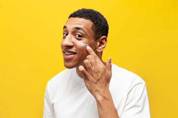 Wall Mural - american guy in white t-shirt applies cream on his face on yellow isolated background, man uses facial cosmetics