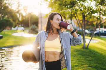 Beautiful Caucasian woman holding a yoga mat in the park drinking a cup of tea or coffee and looking away. Healthy lifestyle, wellness. Healthy lifestyle, sport