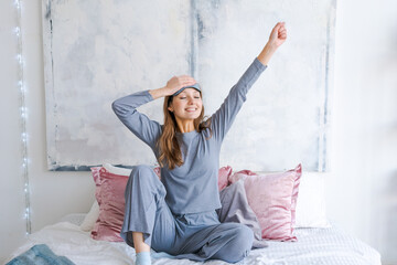 Happy young woman sitting in comfortable bed, arms outstretched after waking up in the bedroom, wearing blue pajamas with a sleep mask, doing exercises in the morning, welcoming a new day, sunrise