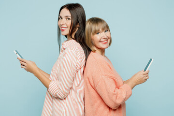 Wall Mural - Sideways elder parent mom with young adult daughter two women together wear casual clothes hold in hand use mobile cell phone stand back to back isolated on plain blue background. Family day concept.