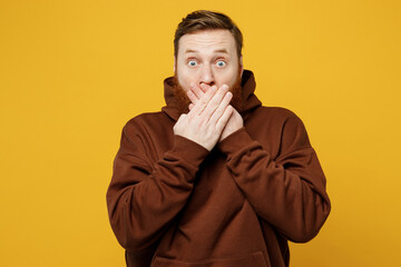 Wall Mural - Young dissatisfied shocked scared sad pop-eyed redhead caucasian man wearing brown hoody casual clothes covering mouth with hand isolated on plain yellow background studio portrait. Lifestyle concept.