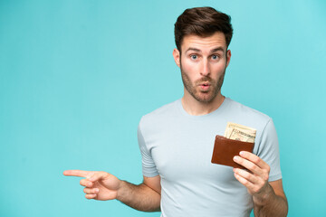 Sticker - Young caucasian man holding a wallet isolated on blue background surprised and pointing side