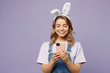 Wall Mural - Young smiling funny woman wearing casual clothes bunny rabbit ears hold in hand use mobile cell phone isolated on plain pastel light purple background studio portrait. Lifestyle Happy Easter concept.