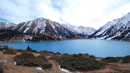 Wall Mural - A mountain lake with blue water in winter. The sun's rays are reflected from the water. Green forest and grass grow on the hills. There are stones lying. The high peaks are covered with snow. Almaty