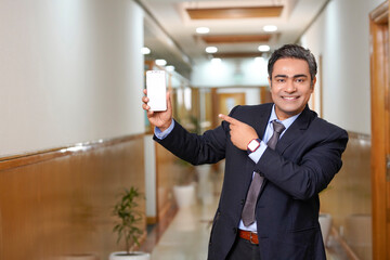 Young businessman showing smartphone screen on office background.