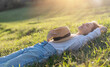 Dreamy young beautiful woman with hat lying on green grass with closed eyes enjoying summer sun and spring day