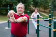 © caftor - Staying after retirement. Happy joyful mature retired sportsman wearing sportswear doing side stretching exercises with arm over his head, exercising outside in city park