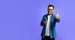 © Moon Safari - Portrait of cheerful young adult man in denim shirt using smart phone and showing thumbs up sign while standing isolated over blue background