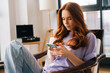 © dikushin - Portrait of thoughtful focused young woman texting message on mobile phone sitting on arm chair. Pensive female using cellphone at home on background of window. Pretty lady chatting online on cell.