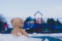 Lonely Teddy Bear Doll Sitting Alone On Footpath With Blurry Kid Playing Playground In Retro Filter, Back View Rear View Lost Brown Bear Toy Looking At People, International Missing Children's Day