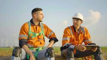 Two Caucasian Engineers Man Worker Wearing Reflective Safety Gear Sit And Rest During Breaks, Chatting, Shake Hand, Exchanging Ideas, Smiling And Having Fun While Sitting In A Row Of Windmill 