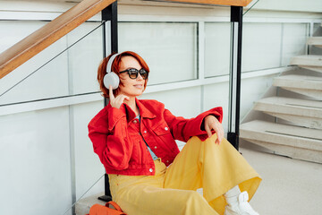 Wall Mural - Smiling hipster young woman in bright clothes, sunglasses with backpack bag and wireless headphones listening to music while sitting on the stairs in public place. Urban city street fashion