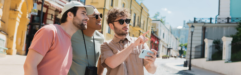Wall Mural - young guide in sunglasses holding smartphone and pointing with finger near multiethnic tourists on a