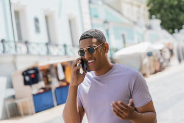 Wall Mural - overjoyed african american man in sunglasses talking on mobile phone on urban street.
