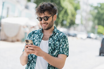 Wall Mural - positive brunette man in sunglasses using mobile phone on blurred urban street.