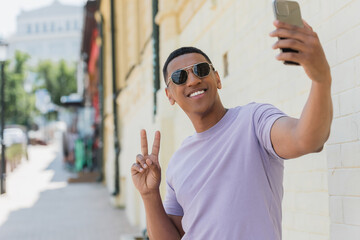 Wall Mural - smiling african american man in sunglasses showing peace sign while taking selfie on blurred urban s