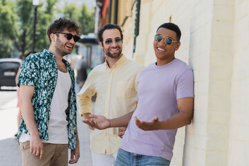 Wall Mural - positive interracial friends in sunglasses standing near building on street in kyiv.