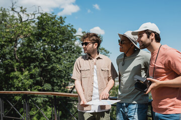 Wall Mural - happy multiethnic travelers in sun hats looking away near tour guide with map in urban park.