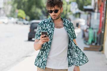 Wall Mural - Brunette man in sunglasses using smartphone while walking on urban street.