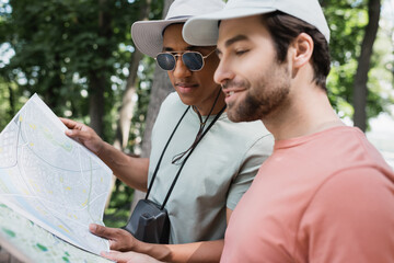 Wall Mural - african american tourist with vintage camera looking at travel map near bearded friend in park.