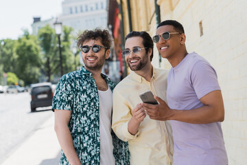 Wall Mural - smiling multiethnic men in sunglasses holding mobile phone and looking away on street in kyiv.
