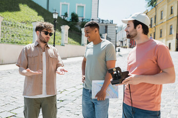 Wall Mural - young tour guide in sunglasses speaking to interracial men with vintage camera and map on andrews de