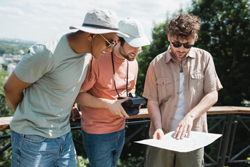 young tour guide in sunglasses showing map to interracial tourists in sun hats during excursion in c