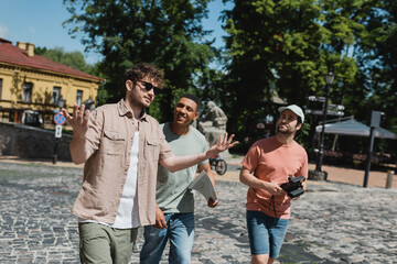 Wall Mural - tour guide in sunglasses gesturing while talking to multicultural tourists during walk on andrews de