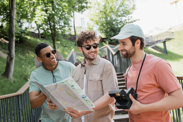 Wall Mural - happy man in sunglasses holding travel map near multicultural friends on stairs of urban street.