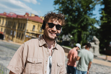 Wall Mural - portrait of curly and carefree tour guide in sunglasses and headset smiling near blurred multiethnic