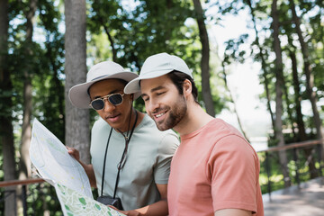 Wall Mural - positive multiethnic tourists in sun hats looking at travel map in blurred park.